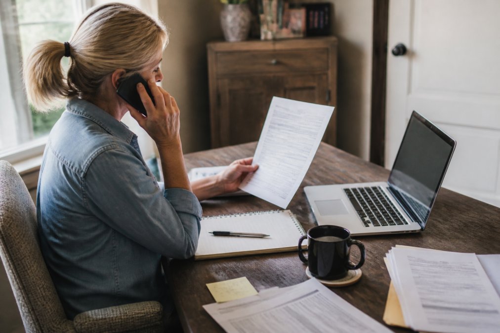 Person on phone call while reviewing insurance documents and health plan information on laptop screen, home office setting with natural window lighting