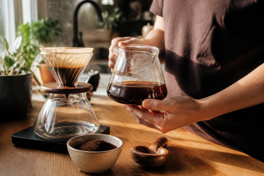 Close-up of hands holding a clear glass pour-over carafe with freshly ground coffee nearby, warm morning light streaming across a kitchen counter