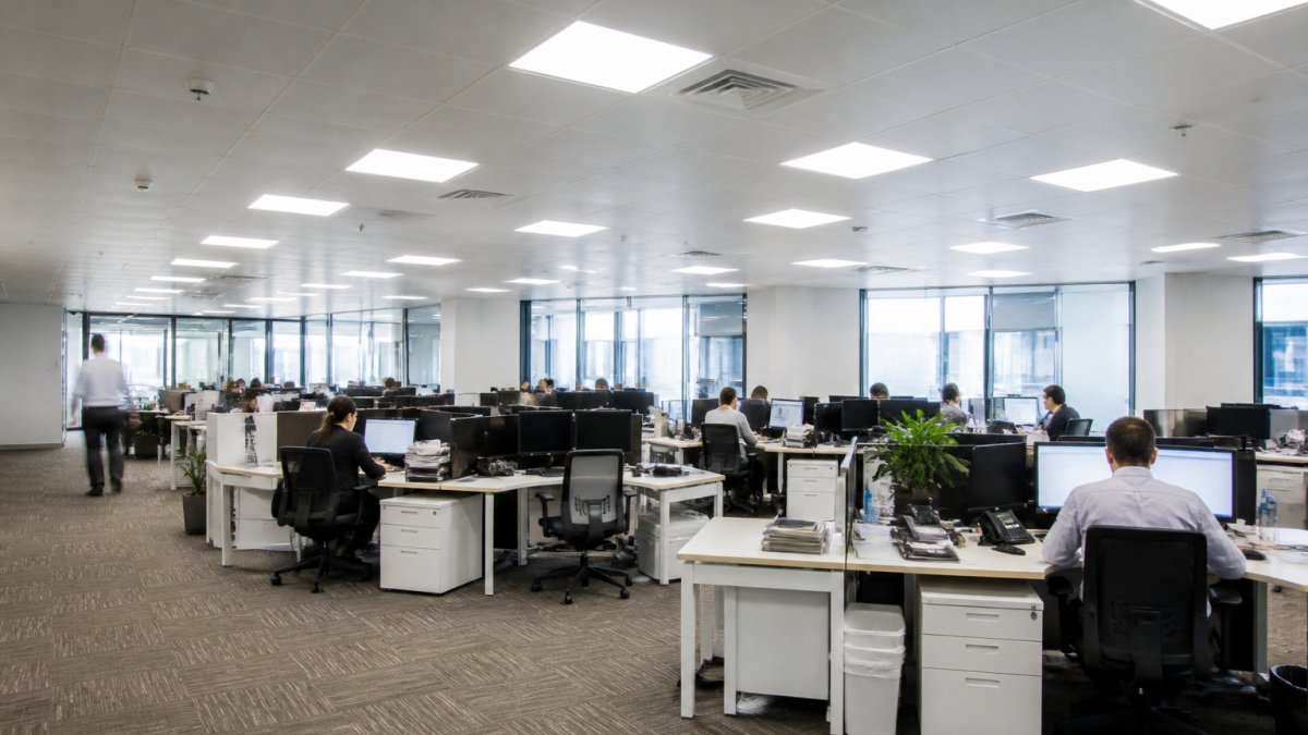 Modern commercial office space with bright LED ceiling fixtures illuminating a well-lit workspace with desks, computers, and employees working efficiently
