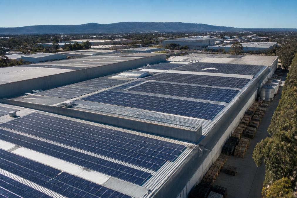 Aerial view of a large manufacturing facility with rooftop solar panels installed across multiple sections of the industrial building, showing the scale of photovoltaic systems mounted on corrugated metal roof surfaces in bright daylight