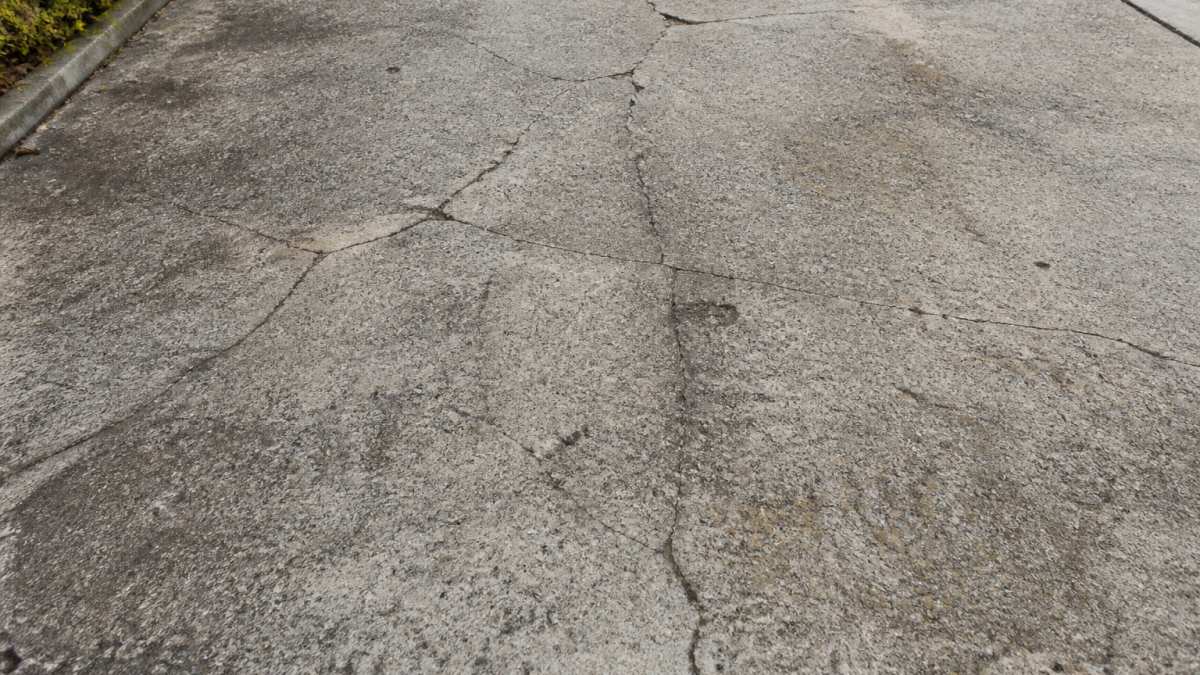 Close-up of a weathered concrete driveway surface showing visible cracks, staining, and wear patterns in natural daylight