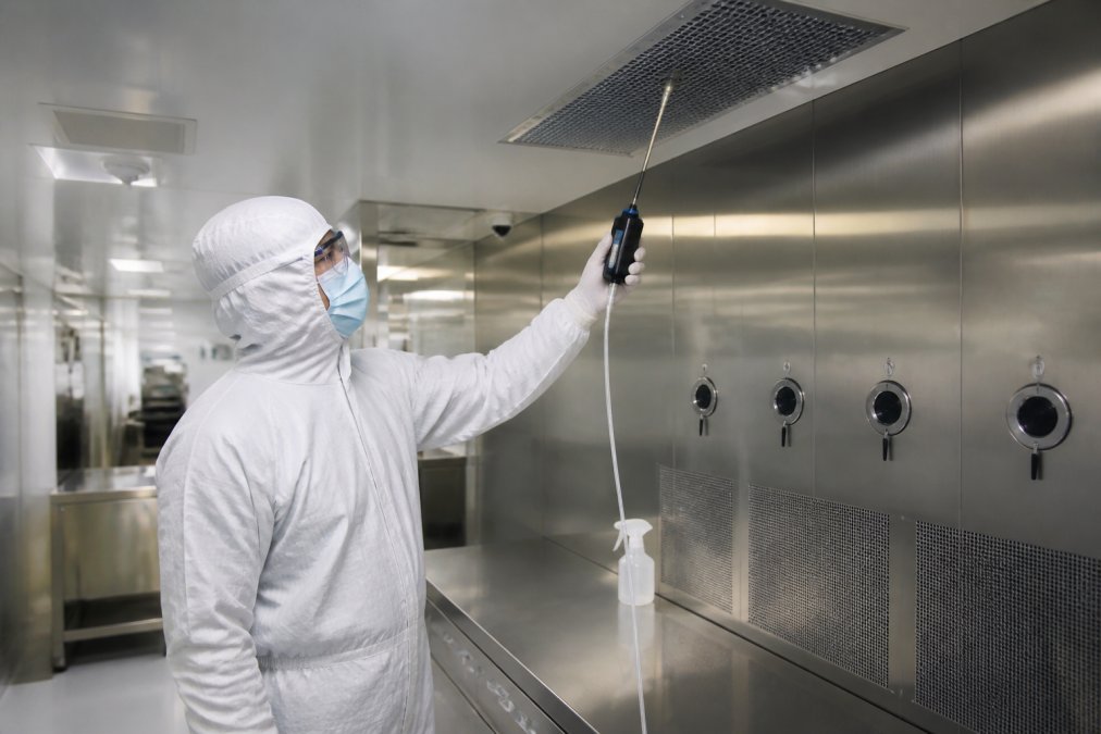 Quality assurance professional in sterile protective gear and full-body gown conducting environmental particle monitoring inside a cleanroom environment with visible HEPA filtration units and stainless steel surfaces