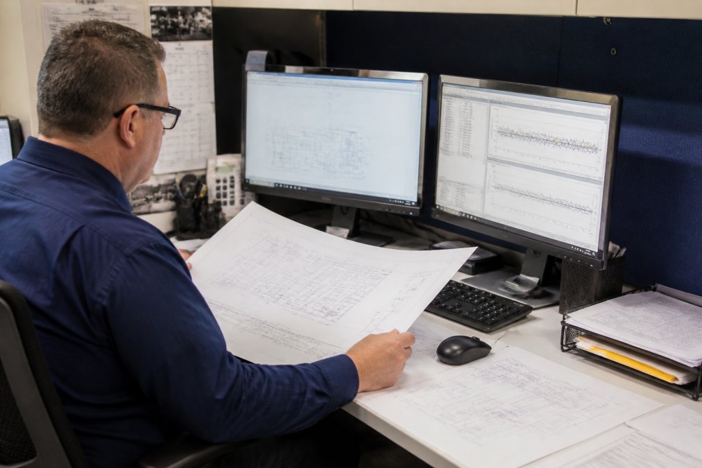 Facilities engineer reviewing detailed cleanroom validation documentation and airflow pattern specifications at a workstation with computer displays showing monitoring data and architectural plans