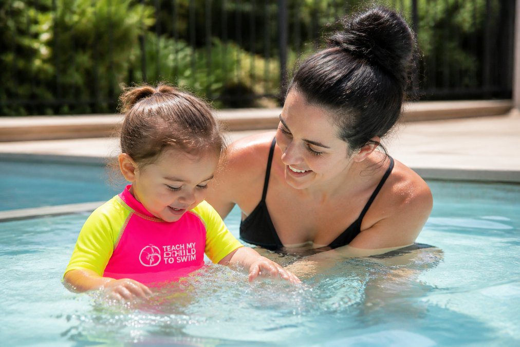 Parent confidently supervising a young child playing in shallow pool water, both smiling and wearing bright-colored swimwear in natural daylight