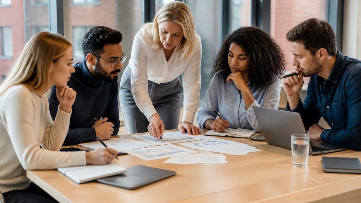 Professional team of diverse colleagues gathered around a modern office table reviewing strategy documents and data sheets, engaged in focused discussion with notepads and laptops, natural daylight from windows illuminating collaborative problem-solving