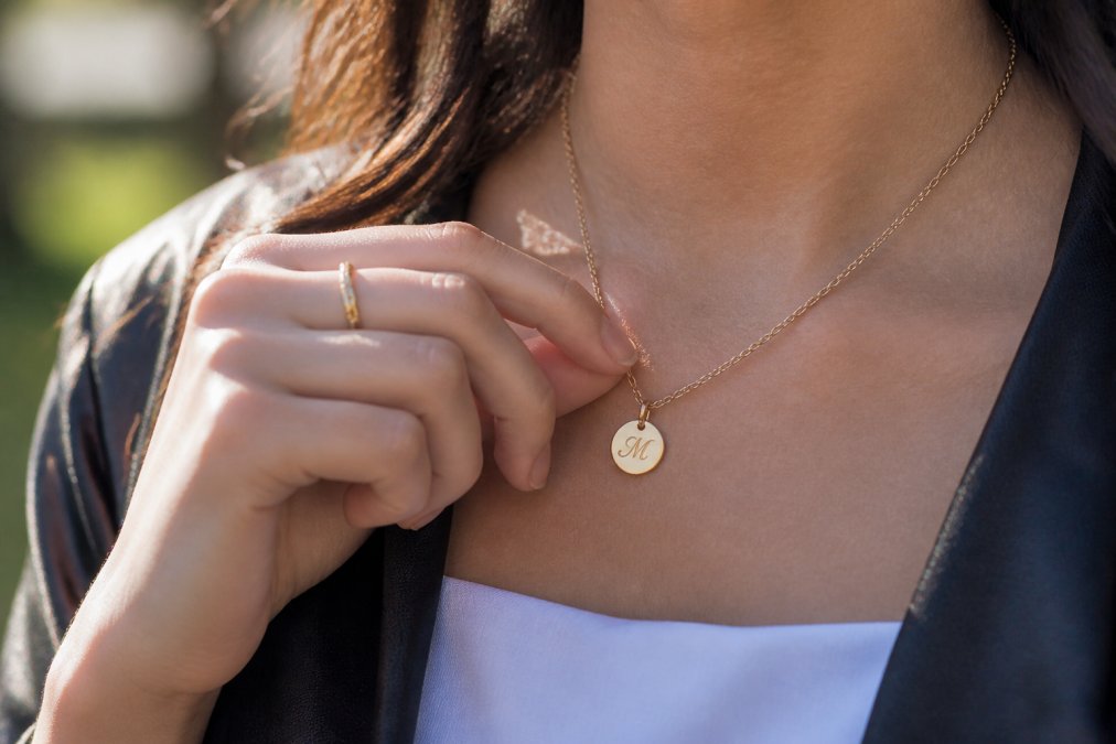 Close-up of a graduate's hand wearing a delicate personalized necklace with engraved initials, showing fine detail against natural sunlight with soft focus background