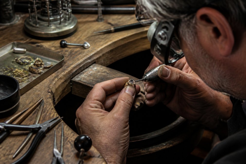 Professional jeweler's hands working on a custom ring at a workbench with precision tools, magnifying glass, and precious metals visible under focused lighting