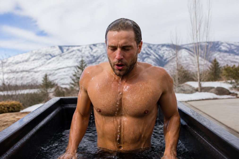 Person emerging from a cold plunge with visible cold shock response, water dripping from face and shoulders, focused expression, outdoor setting with modern cold plunge visible in background
