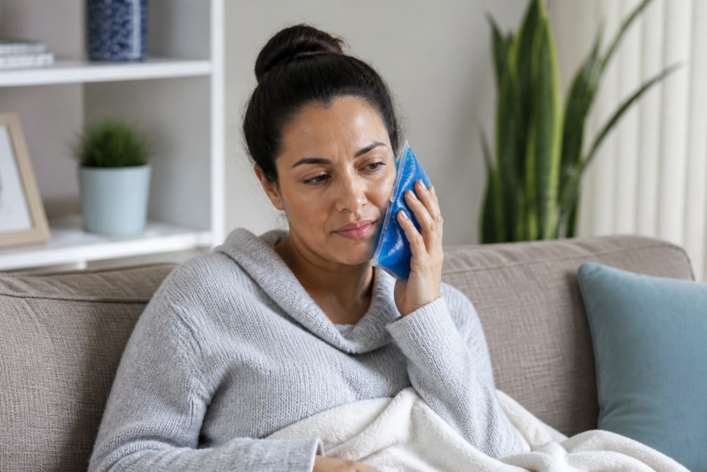 Person at home applying ice pack to cheek with gentle expression, demonstrating post-surgical pain management technique in comfortable home setting
