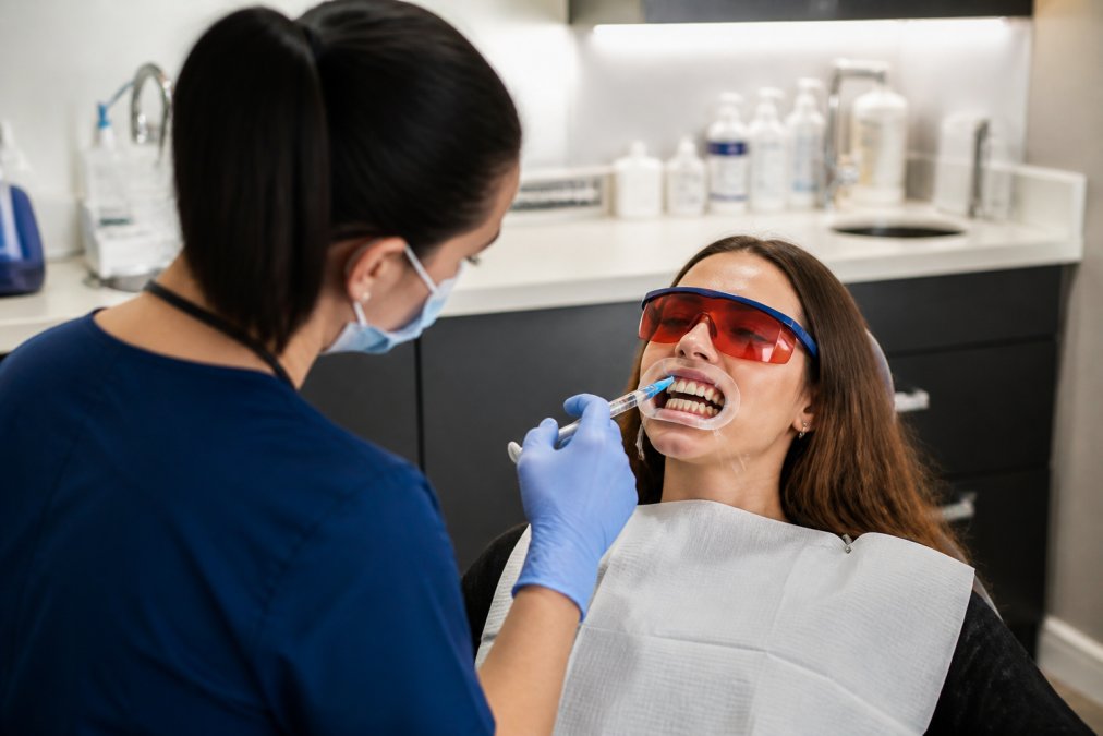 Dentist carefully applying professional whitening gel to patient's teeth during in-chair treatment, with patient relaxed in modern dental chair under soft clinical lighting