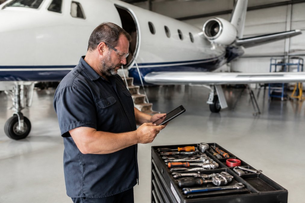 Aircraft maintenance technician reviewing digital logbook records on tablet in aircraft hangar with tools and aircraft visible in background, natural hangar lighting