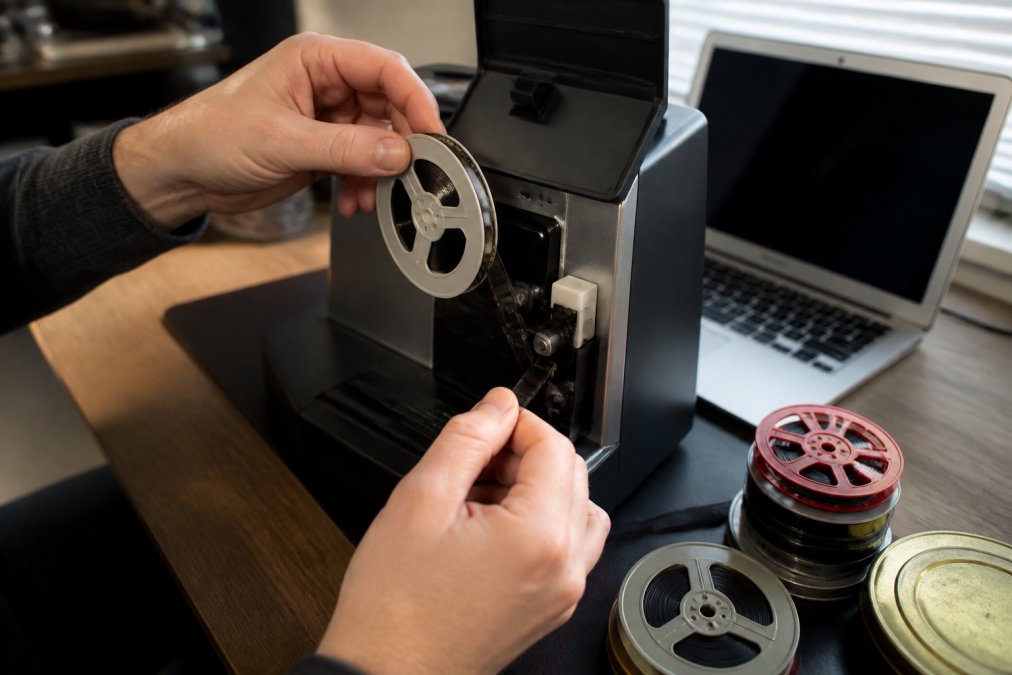 Close-up of a person's hands threading a small 8mm film reel onto a home scanner in a well-lit home office, with other reels stacked nearby and a laptop showing a scanning interface