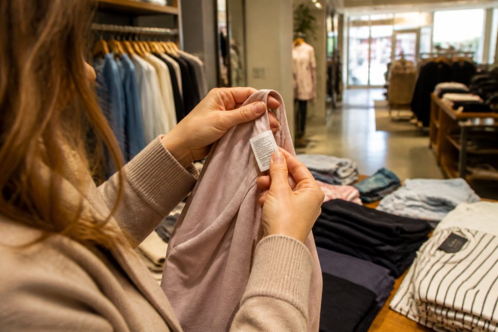 Shopper carefully examining clothing tags and fabric content labels in a bright retail environment, hands holding garment up to light while reading fiber composition