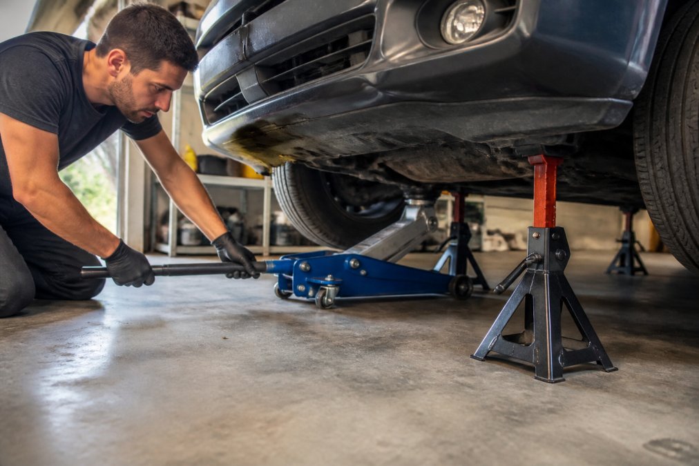 A person safely lifting a vehicle with a floor jack while jack stands are positioned underneath, showing proper safety setup in a well-lit garage with concrete floor