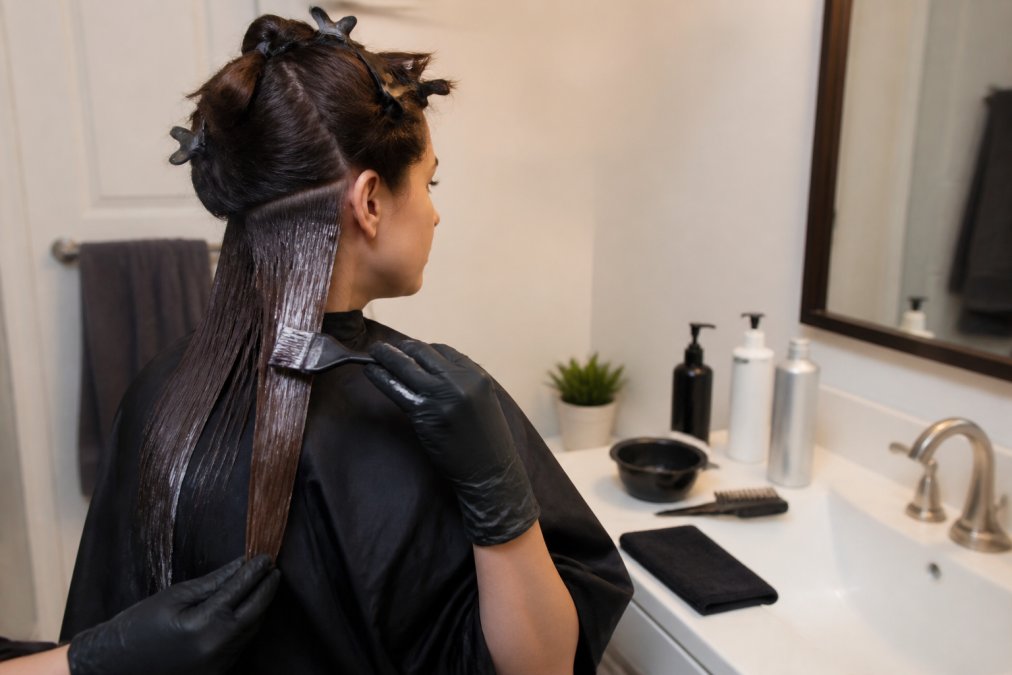 Person applying keratin treatment to sectioned hair at home, wearing protective gloves, with product bottles and supplies arranged on bathroom counter near sink