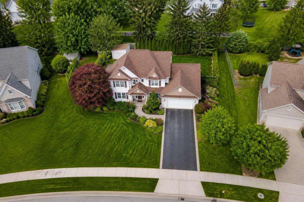 Aerial drone shot of a suburban home with manicured lawn, driveway, and surrounding neighborhood visible from above, showing property boundaries, mature trees, and adjacent properties in clear detail