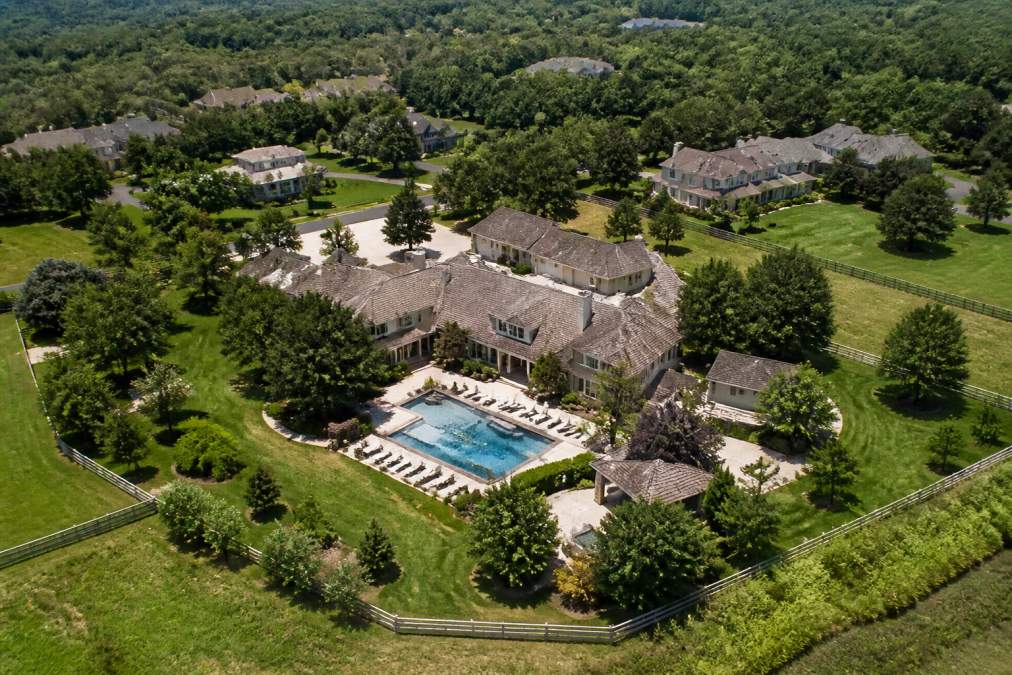Overhead drone photograph of a luxury residential estate showing a large main house with attached structures, swimming pool with surrounding patio, mature landscaping, extensive grounds, and adjacent properties visible in the distance