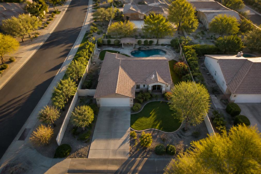 Aerial view of a small residential property showing the full lot, landscaping, driveway, and surrounding neighborhood context from above, shot during golden hour with clear shadows defining the landscape