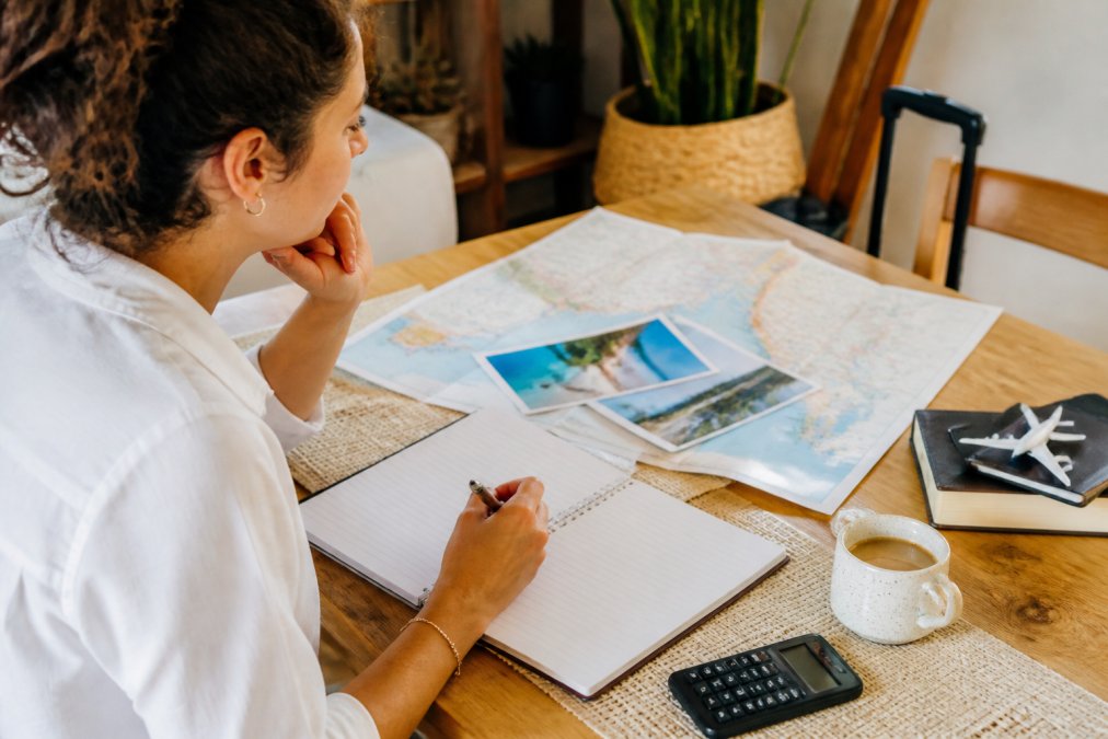 Iemand die op een laptop vakantiebestemmingen onderzoekt met een notitieboek en koffie, terwijl ze hun reisbudget plannen