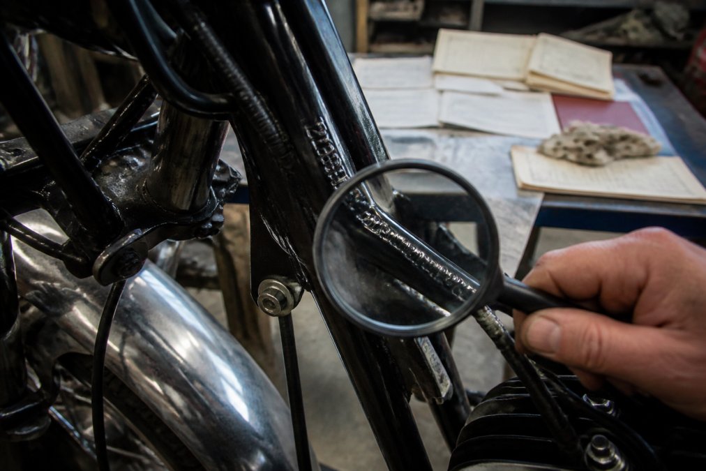 Close-up van een origineel chassisnummer op een klassiek motorfietsframe, met een vergrootglas en onderhoudsdocumenten op een werkbank in een goed verlichte garage