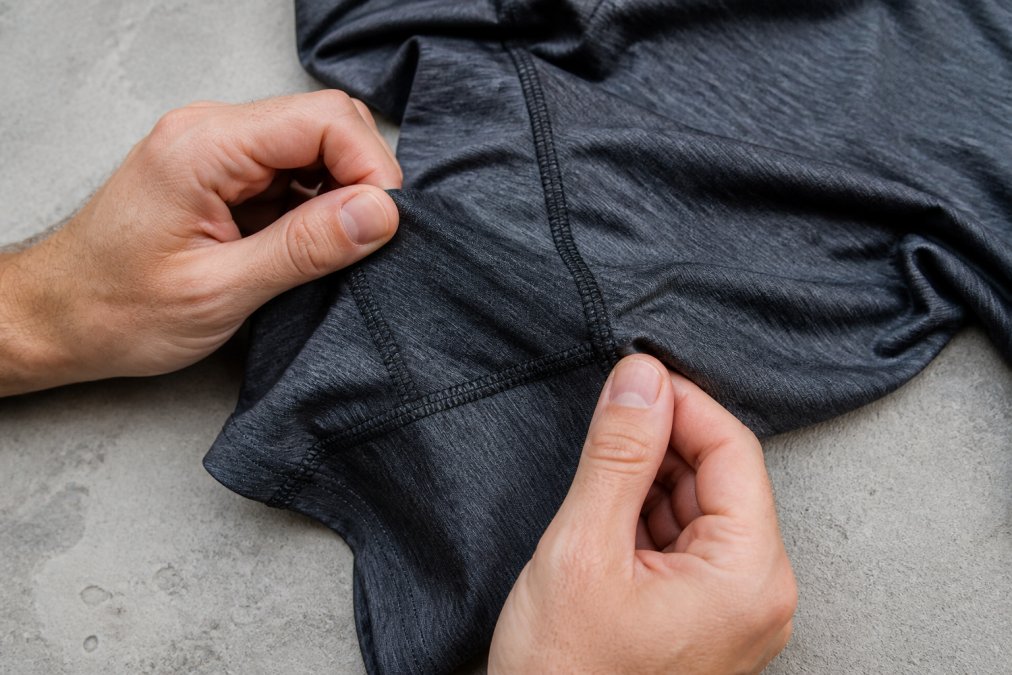 Close-up of hands examining the stitching and fabric of a high-performance athletic shirt, showing flatlock seams and synthetic blend material in natural light