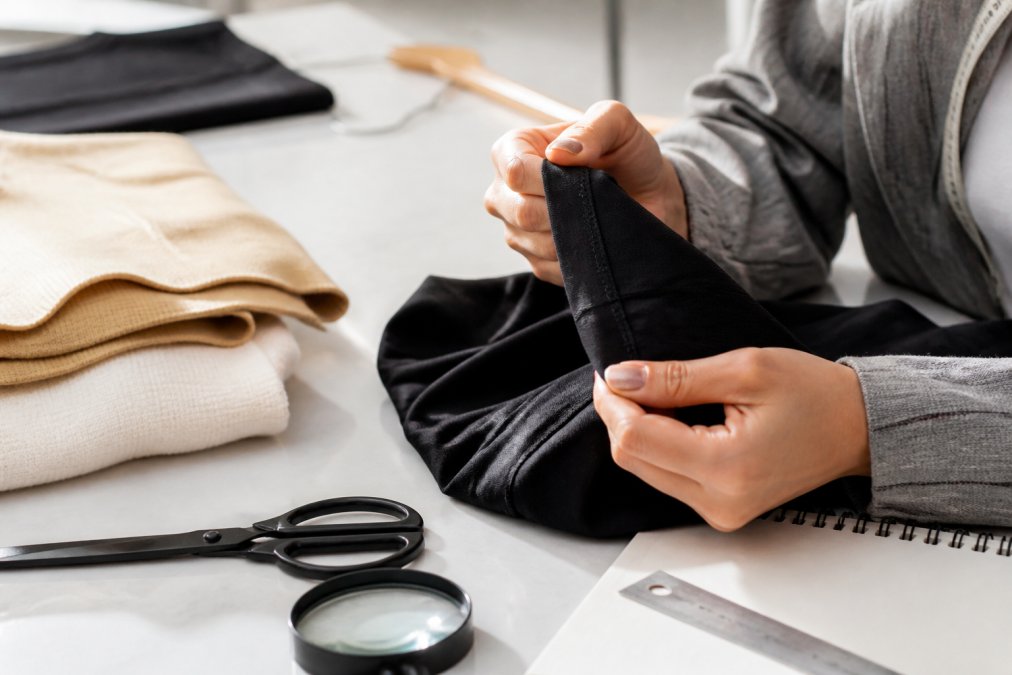 Close-up of hands examining a garment's seams, stitching details, and fabric texture against a neutral background with natural daylight illuminating the inspection