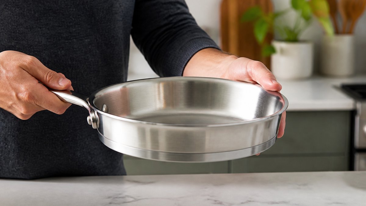 Close-up of hands holding a premium stainless steel pan, showing the substantial weight and layered construction, with a modern kitchen countertop in soft natural lighting