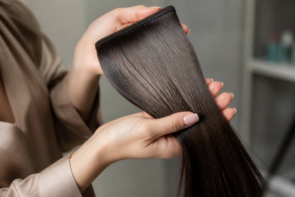 Close-up of a woman's hands holding a section of premium human hair extensions against a soft neutral background, showing natural shine and texture