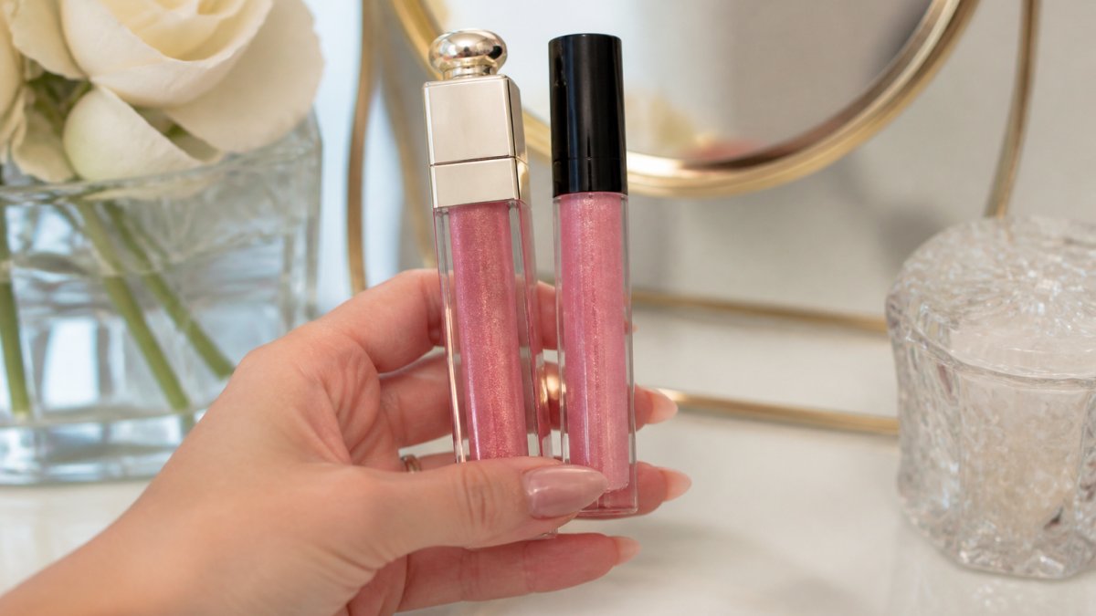 Close-up of a woman's hand holding two lip gloss tubes side by side, one sleek and one basic, with a soft focus on a vanity mirror in the background