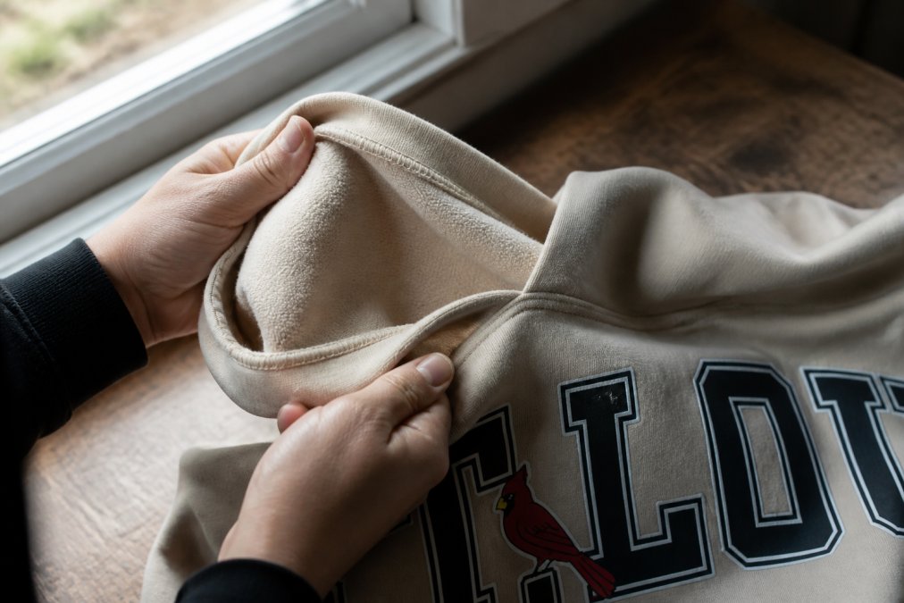 Close-up of hands examining the interior seams and fabric weave of a premium cotton hoodie under natural window light, showing stitch density and material texture in detail