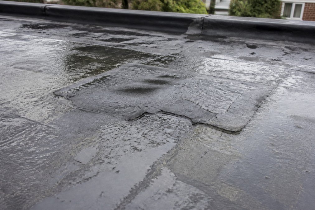 Close-up of a flat roof showing visible blistering, cracking, and water damage around a previous patch repair, with moisture visible on the membrane surface