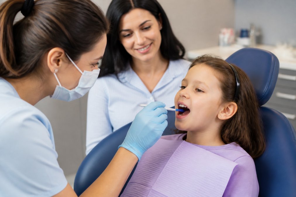 Pediatric dentist applying fluoride treatment to a young child’s teeth while a parent watches in a modern clinic