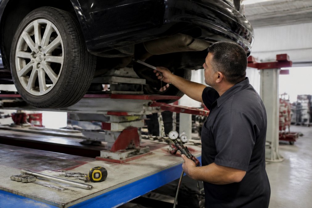 A certified technician in a clean auto body shop examining the underside of an older sedan on a frame machine, with measuring tools visible nearby