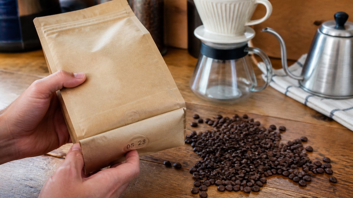 Hands holding a coffee bag with a roast date next to whole beans, a pour-over dripper, and a gooseneck kettle.