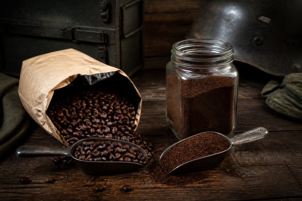 A side-by-side comparison of coffee beans: one bag of whole beans in a vintage-style package next to a glass container of pre-ground coffee, with a scoop of each on a dark wood surface