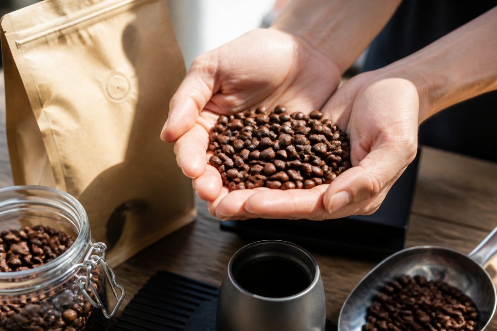 Close-up of hands holding freshly roasted coffee beans with vibrant brown color and subtle surface sheen, roast date label visible on premium coffee bag in natural sunlight