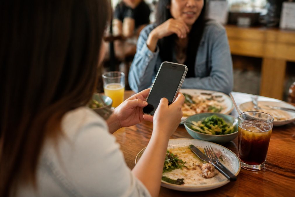 A young diner at a casual restaurant table holding a smartphone showing a rewards screen, with a half-eaten meal and a friend across the table, warm natural light