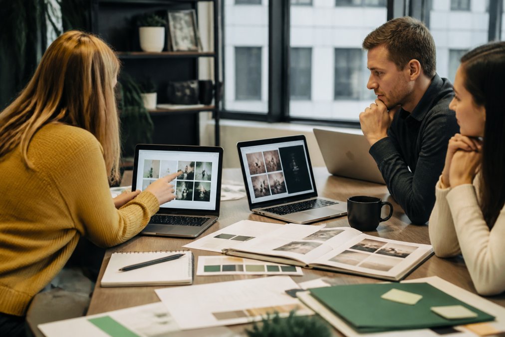 A marketing team reviewing AI-generated content on multiple laptop screens in a bright modern office, with brand style guides and printed notes spread across the desk