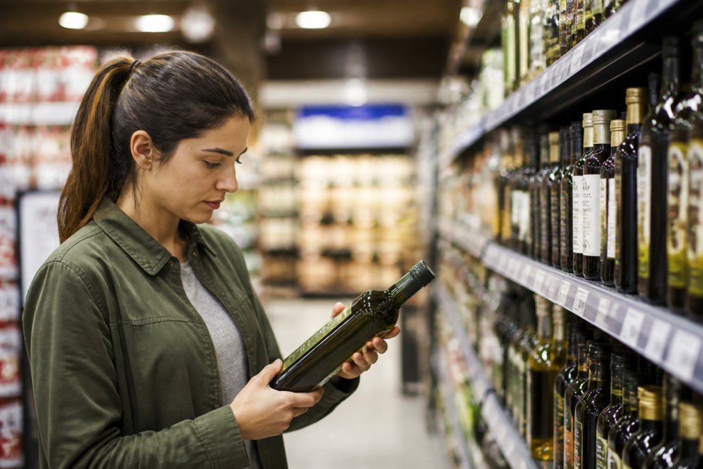 Een persoon in een supermarkt die een fles olijfolie vasthoudt en het etiket leest, met de focus op de oogstdatum en herkomst, fel winkellicht