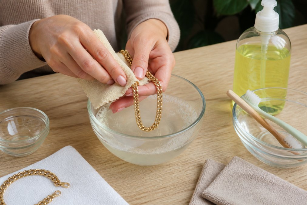 Hands gently cleaning a gold plated necklace with a soft microfiber cloth over a small bowl of mild soapy water, with a bottle of gentle dish soap and additional cleaning supplies visible on a light wooden surface