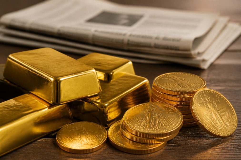 Close-up of several gold bullion bars and gold coins arranged on a dark wooden surface, with a folded financial newspaper visible in the soft-focus background, warm studio lighting
