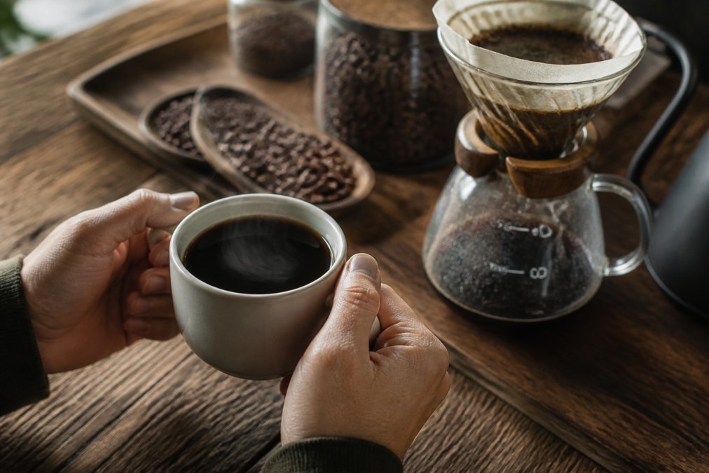 Hands holding a steaming ceramic cup of coffee with a pour-over setup and fresh beans on a wooden counter