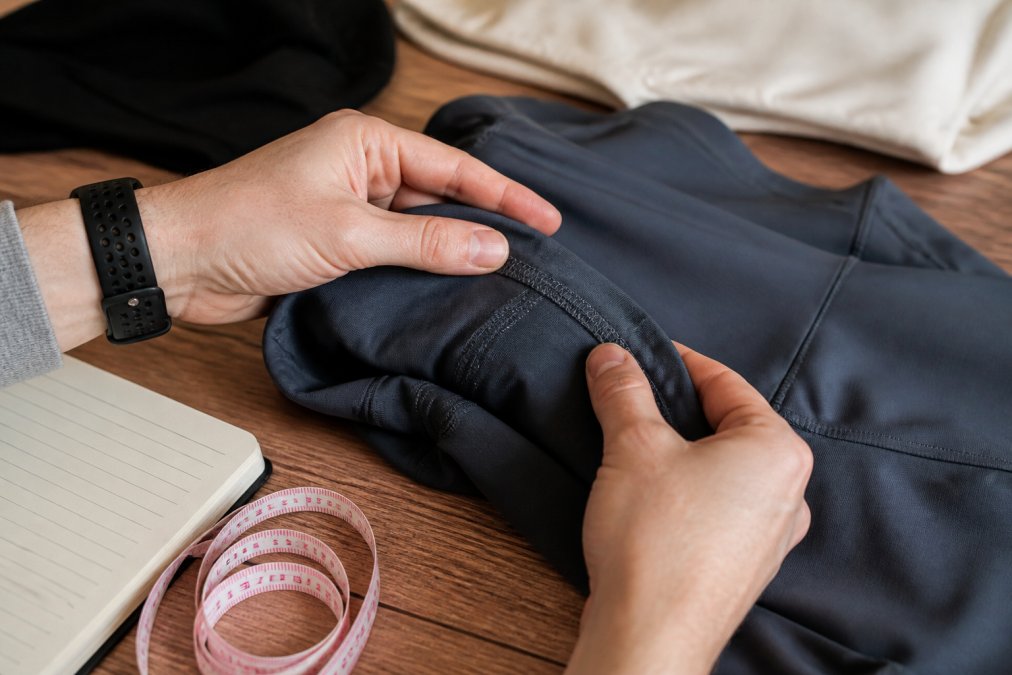 Close-up of hands examining the seams and stitching on a folded piece of athletic clothing on a wooden table, with a soft measuring tape and notebook beside it