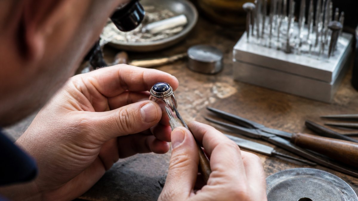 Close-up of jeweler's hands examining a sterling silver ring with sodalite stone under bright studio lighting, using magnification tools to inspect detail work and gemstone setting