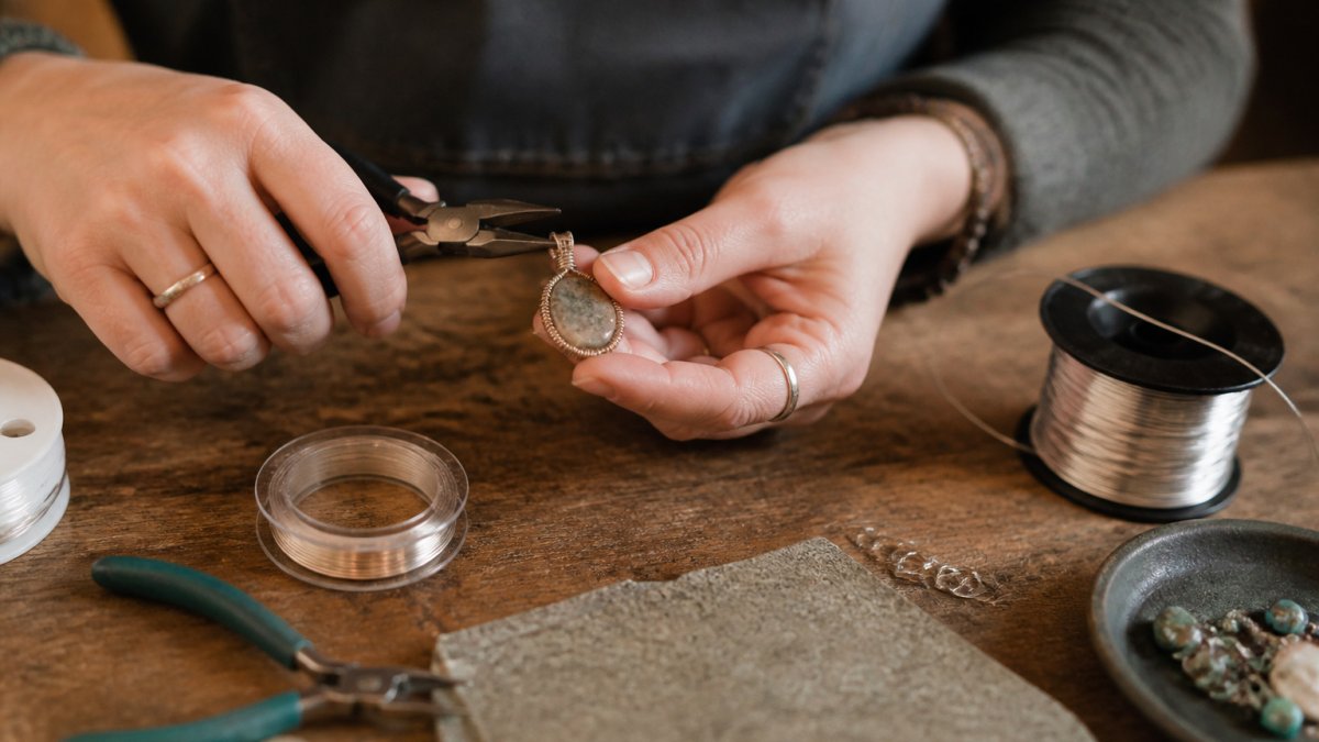 Jewelry maker using pliers to wire-wrap a gemstone pendant at a wooden workbench with silver wire spools