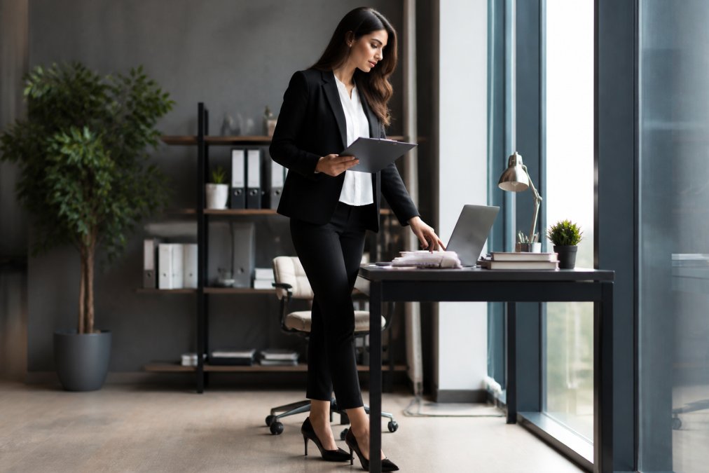 Professional woman in business attire wearing high heels, standing confidently at a modern office desk with laptop and documents, appearing poised and comfortable