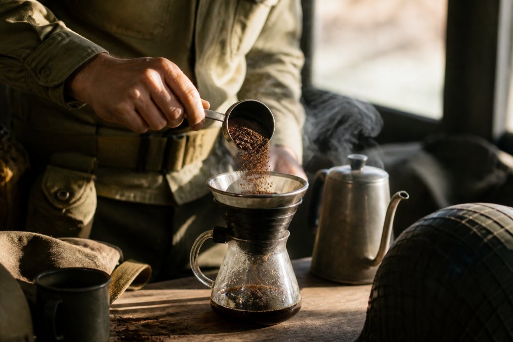 Close-up of freshly ground dark roast coffee being measured with precision into a brewing vessel, steam rising from hot water, showing the exact moment grounds meet water in morning light