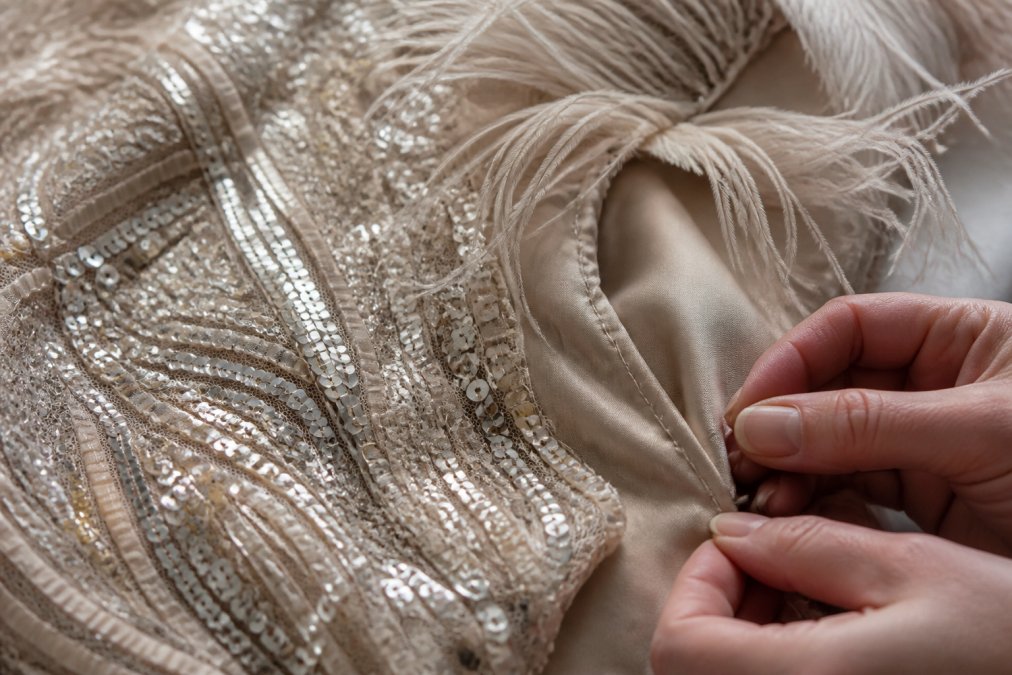 Close-up of intricate sequin embellishment, delicate feather trim, and precision stitching on luxury evening fabric, showing craftsmanship detail under soft lighting
