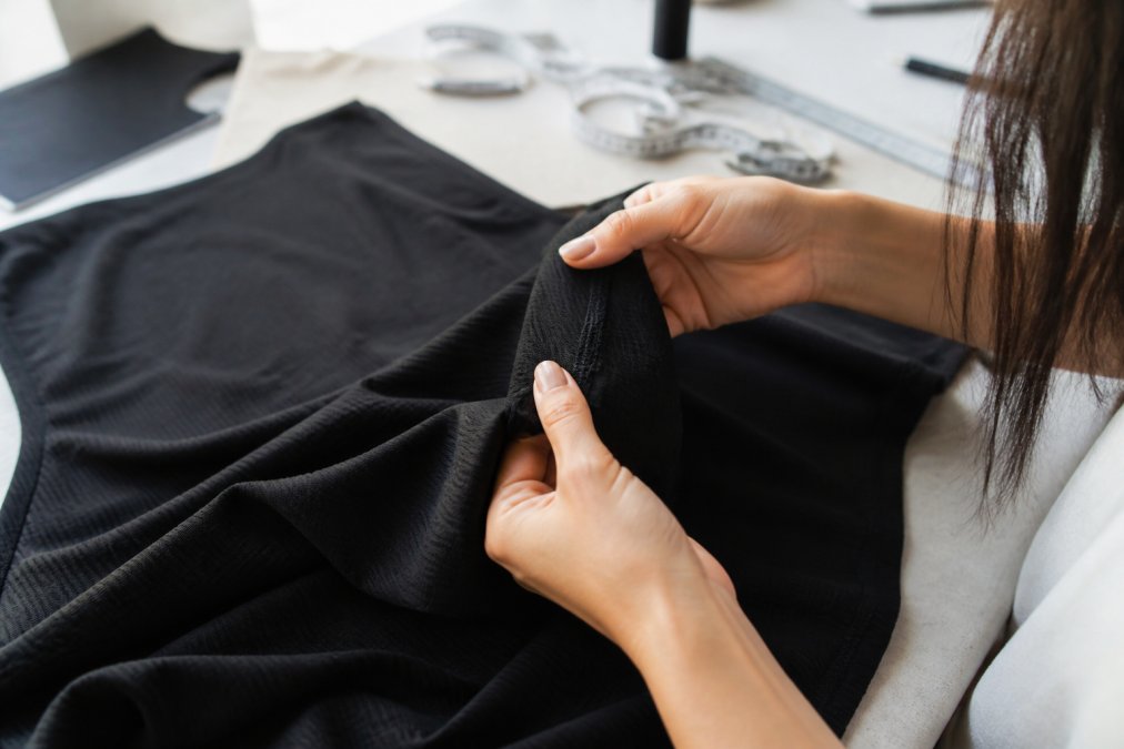 Close-up of a woman's hands gently examining the fabric and seam of a dark knit maxi dress, with soft natural light highlighting the texture and stitching detail