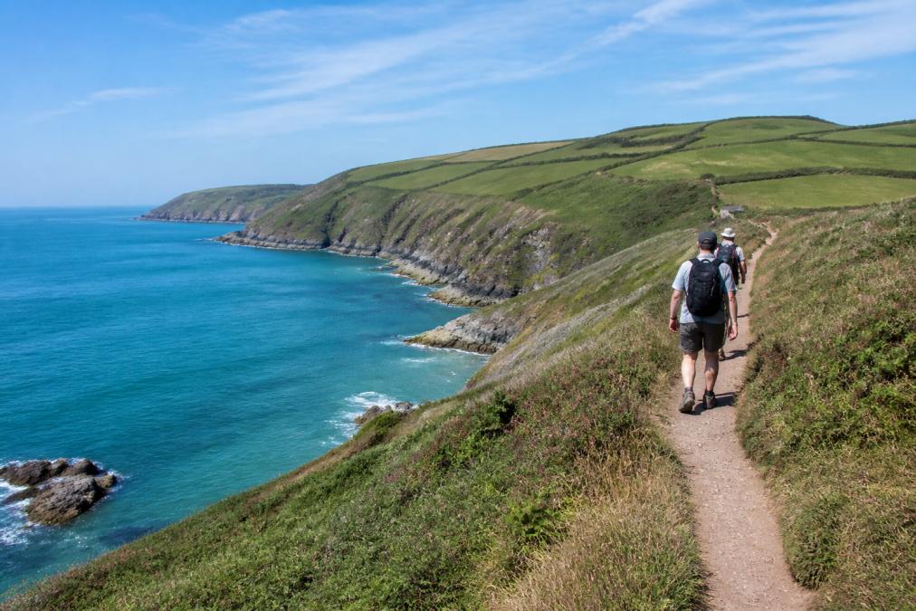 Hikers walking along a dramatic clifftop coastal path with turquoise sea views below, green hillsides rolling inland, and a clear blue sky stretching across the horizon